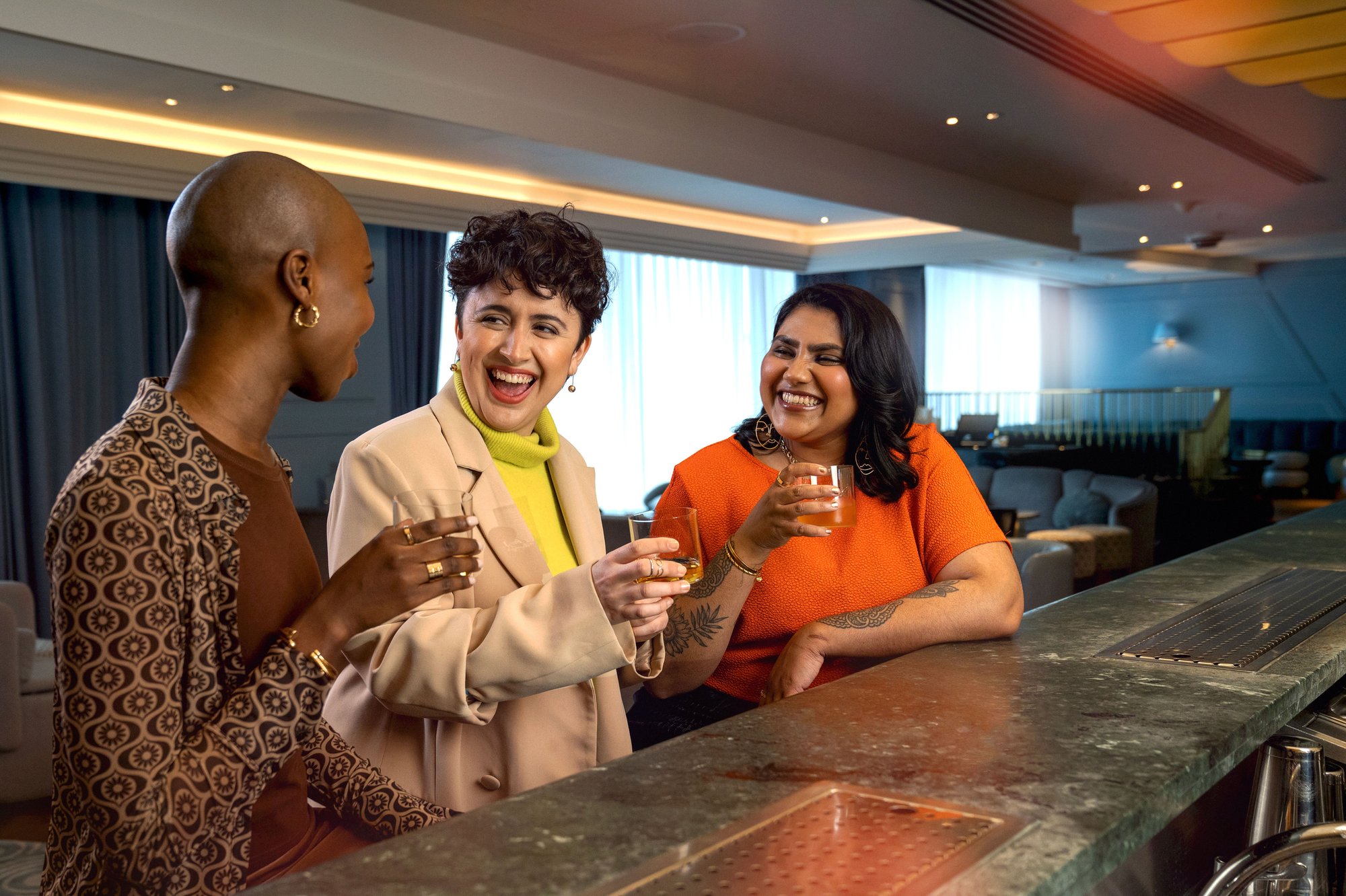Three women laughing over drinks at a bar counter